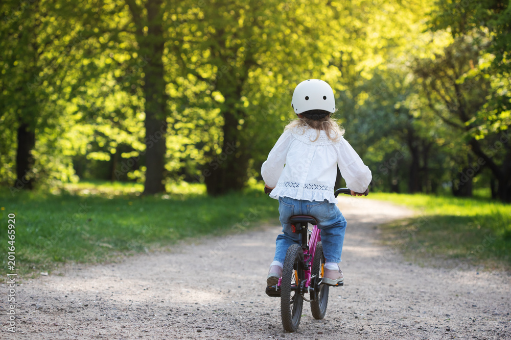Fototapeta premium Little girl in cycling helmet enjoying riding bicycle in nature outdoor, toddler active leisure, happy bike ride on park trail in sunlight.