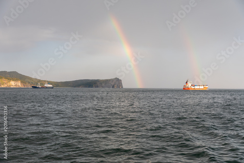 Wide seascape with cargo ships and a double rainbow over Avacha Bay near Petropavlovsk-Kamchatsky, Kamchatka Krai, Russia, with cliffs and calm water