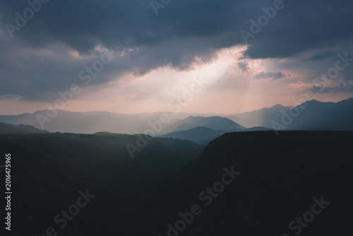 Wide landscape view of layered mountain ridges and a deep canyon at sunset in Durmitor National Park, Montenegro, with sun rays breaking through dark storm clouds