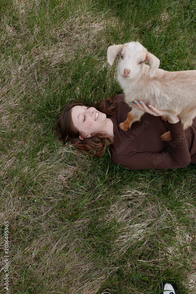 Obraz premium Casual lifestyle scene shows a woman lying on grass outdoors, a young goat kid perched on her chest, sharing an affectionate moment amid meadow, showcasing simple joy and bond with animals in a
