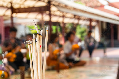 People sit in a temple as incense sticks burn in the foreground during a daytime gathering for prayers and rituals
