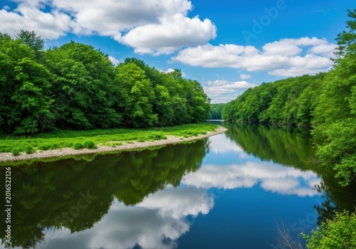 A peaceful, meandering river snakes through a vibrant green woodland, reflecting the clear blue sky and fluffy white clouds above the water surface, stream, bank, current