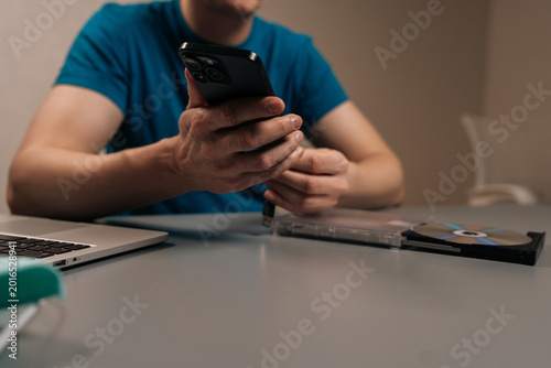 Cropped shot of man connecting smartphone to external optical disc drive on a desk beside laptop, showing a media disc and glowing blue usb ports for data transfer and hybrid work setups.