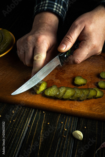 A pair of hands holds a knife and cuts pickles on a wooden cutting board. The scene takes place in a kitchen, conveying an everyday cooking activity