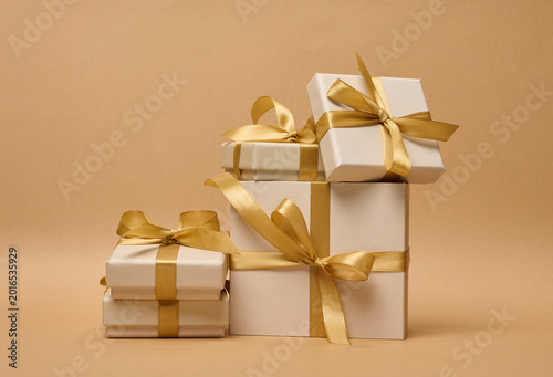 A stack of cream-colored gift boxes tied with gold satin ribbons and bows on a beige background.