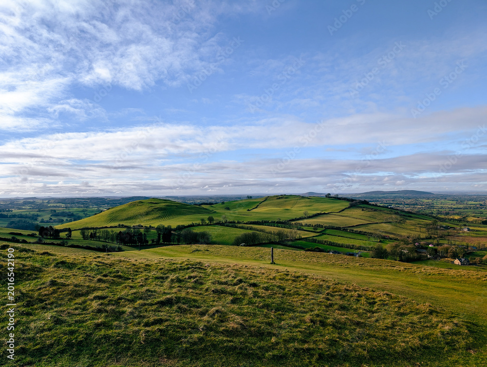 Obraz premium Rolling green hills and fields stretch across the Irish countryside under a bright blue sky with soft clouds