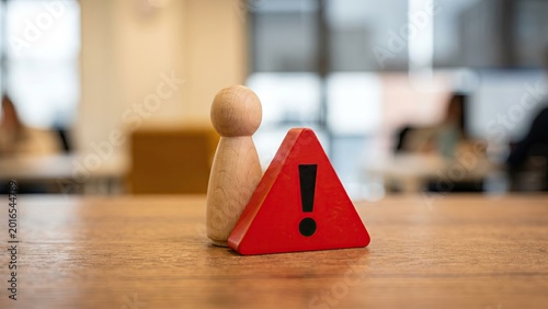 A wooden peg figure standing behind a small red warning triangle on a desk, symbolizing crisis management, risk alert and business continuity.