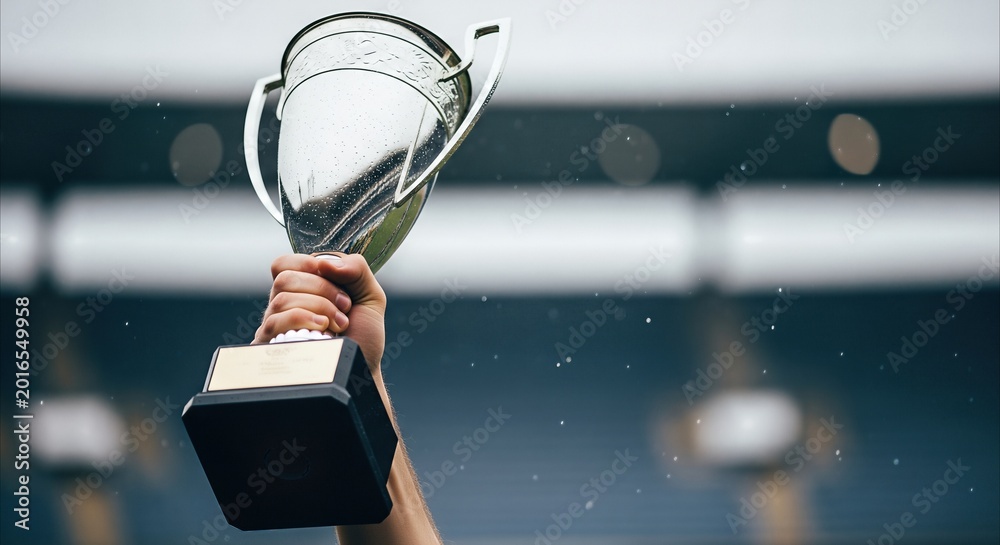 Fototapeta premium Hand holding a silver trophy aloft against a stadium background, symbolizing victory and achievement in a dramatic, cinematic style with copy space