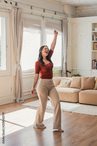 Happy young woman joyfully dancing and singing with raised arm, feeling freedom and positive emotions while enjoying music in her bright living room at home