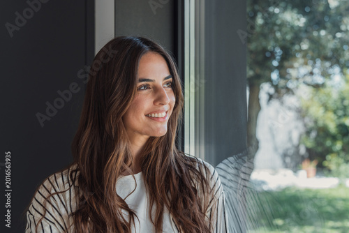 Photos Young woman smiling while looking out a large window, contemplating the future w