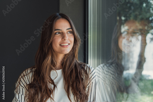 Quadro su tela Young woman smiling with thoughtful expression while looking out a large window,