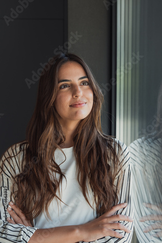 Young woman with long brown hair stands by a window with arms crossed, smiling softly and looking up, daydreaming about future possibilities with calm confidence and peace of mind
