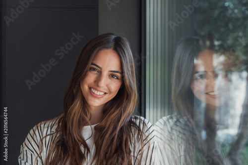 Tableau sur toile Young woman smiling by a window with her reflection visible, radiating warmth, c