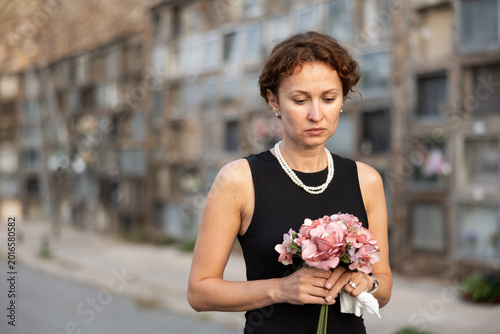 Bild auf Leinwand Sorrowful middle aged woman in black dress standing in cemetery holding bouquet