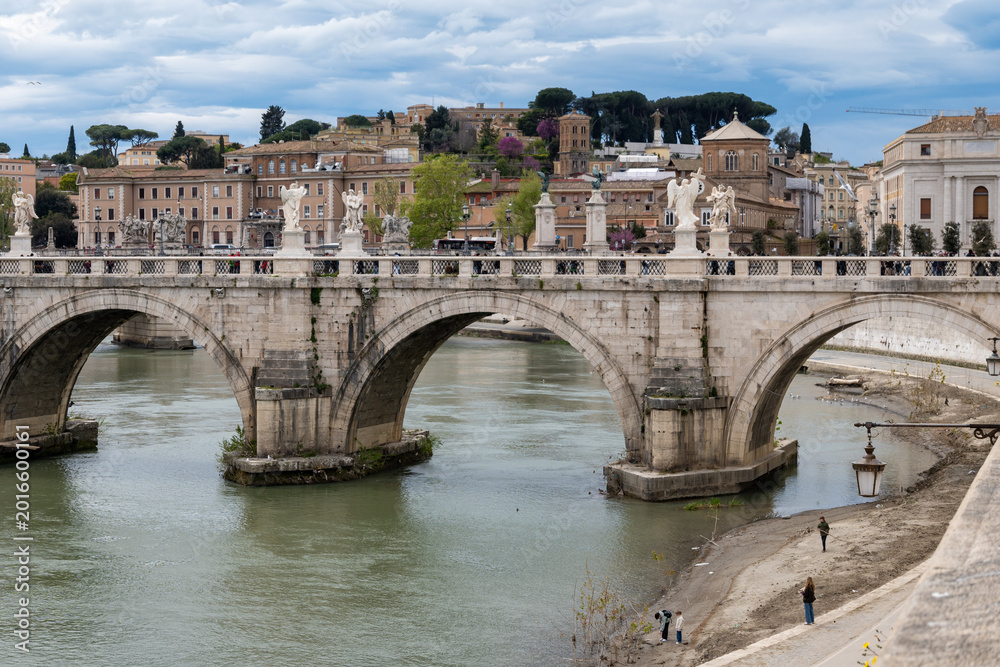 Fototapeta premium View on stone-arched bridge on Tiber river in Rome, balustrade, marble statues, terracotta buildings, domes and umbrella pines rise on gently sloping Vatican hill, classic Roman skyline