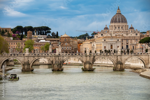 View on stone-arched bridge on Tiber river and St Peter's Basilica in Rome, terracotta buildings, domes and umbrella pines rise on gently sloping Vatican hill, classic Roman skyline