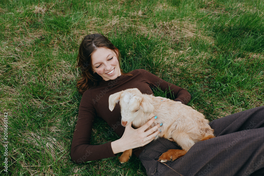 Fototapeta premium Happy woman lying on grass beside a playful goat kid, embracing nature and friendship in an outdoor lifestyle moment, warm sunlight, relaxed mood, gentle bonding between human and animal