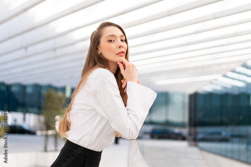 Fototapeta premium Young businesswoman in white shirt posing thoughtfully near modern office building with glass facade and architecture, showing confident personality