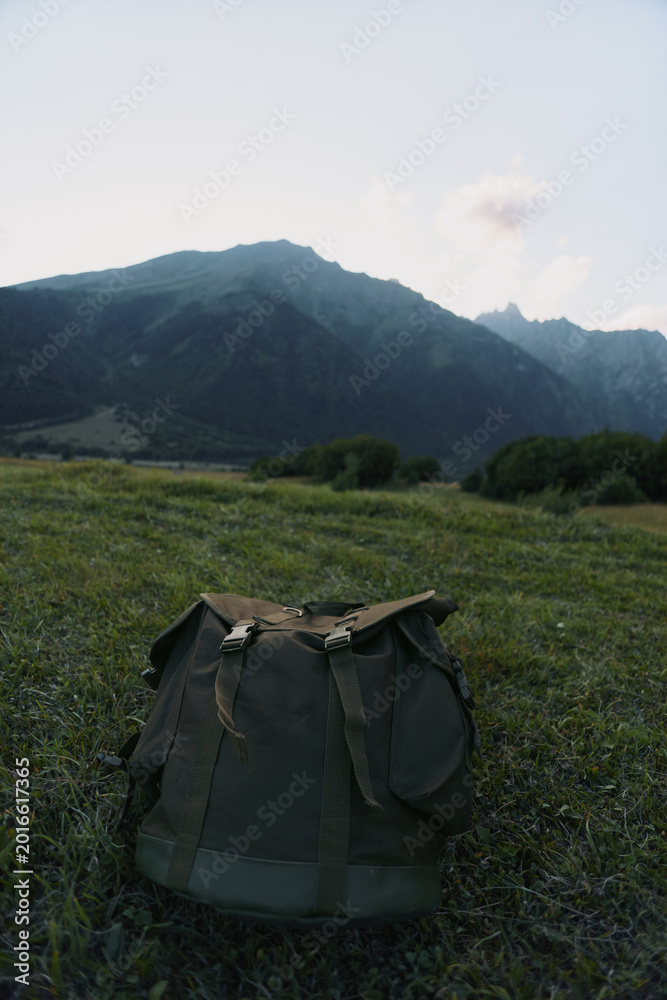 Obraz premium Rugged backpack resting in a grassy field with distant mountains, an outdoor lifestyle scene that conveys adventure, solitude, and simple travel gear in natural light