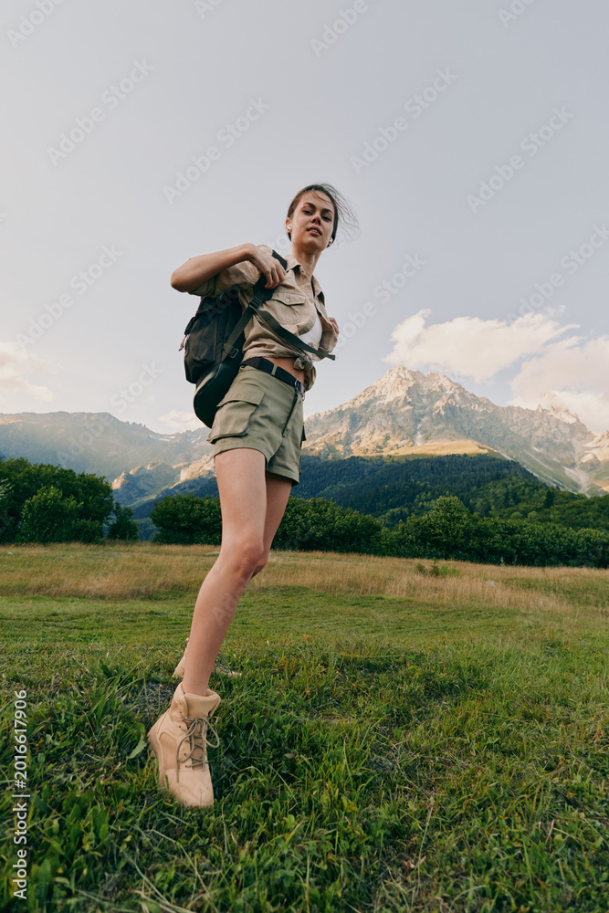 Obraz premium Young woman with a backpack leaps across a grassy field as mountains rise behind, capturing a lively outdoor hiking moment under pale blue sky with soft light and a gentle breeze gently.