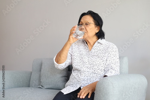 Relaxed calm Asian elder retired woman drinking a glass of natural fresh water, keeping healthy nutrition, diet, hydration balance for dry skin care, metabolism
