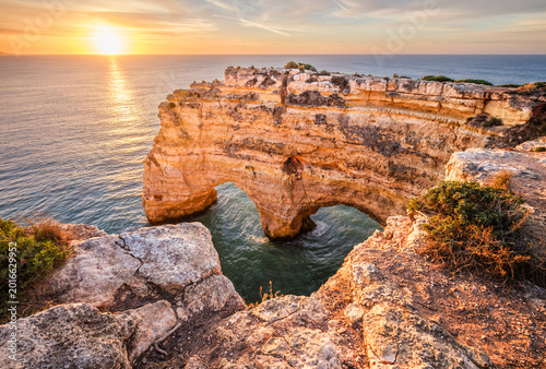 Sunrise view of Praia da Marinha beach in Algarve, Portugal, with a limestone sea arch, Atlantic water, warm sky, and rugged coastal cliffs.