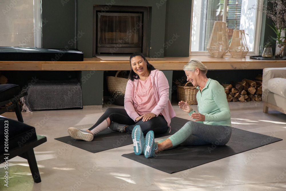 Obraz premium Diverse senior women sitting on black exercise mats in living room, laughing in athletic clothing