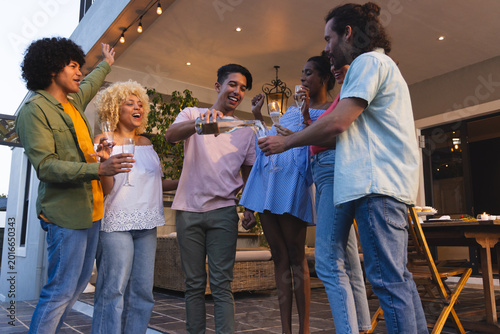 Diverse group raising glasses and pouring from champagne bottle on covered patio with string lights