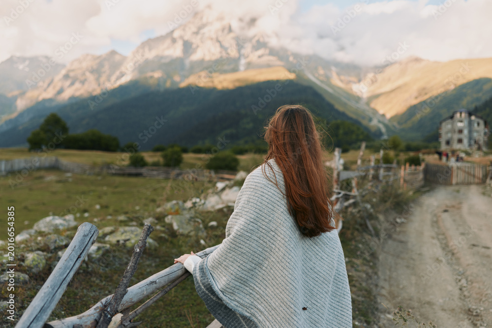 Naklejka premium Outside scene with a woman seen from behind, wearing a cozy cardigan, gazing toward distant mountains, a relaxed lifestyle moment in natural light.
