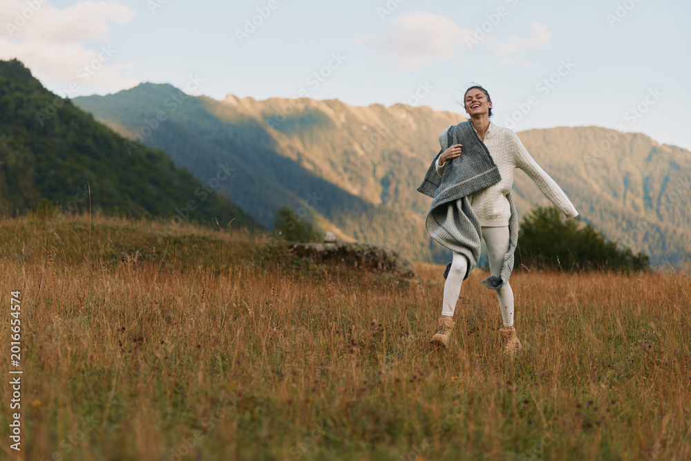Naklejka premium Young woman with long hair joyfully runs through a grassy field in casual clothes during golden hour with mountains in the background and a relaxed expression on her face.