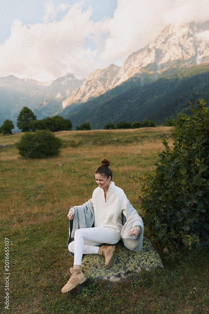 Naklejka premium Woman outdoors in a mountain meadow, wearing a cozy sweater, sitting on a rock, relaxed pose, soft natural light, enjoying the fresh breeze, lifestyle moment