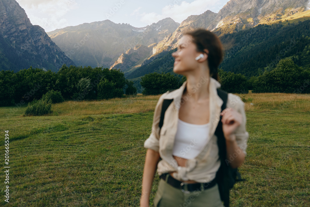Fototapeta premium Young woman with short hair enjoys a mountain hike wearing casual clothes and earbuds while standing in a green meadow with forest and peaks in the background.