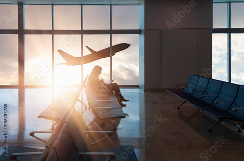 Silhouette of young woman working on laptop and waiting for flight in the Airport - Passenger airplane take off behind airport window at sunset
