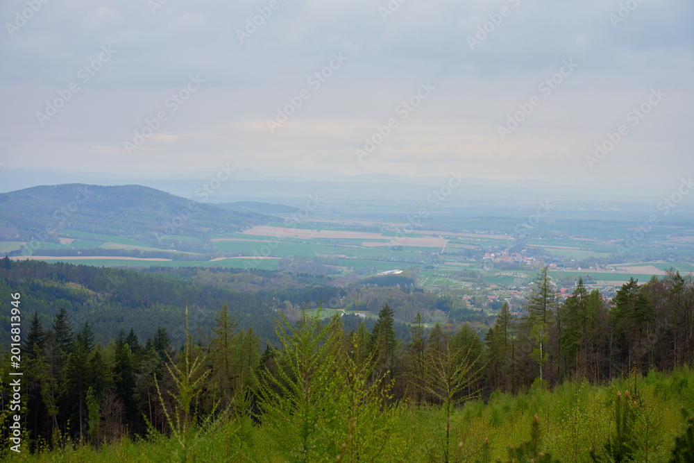 Obraz premium Wide view from a mountain slope over green forest, rolling hills, farmland and small villages in a misty valley under cloudy sky. Nature, rural landscape, travel, countryside and environment concepts
