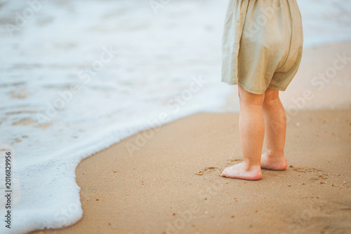 Little Asian toddler girl standing barefoot on a sandy beach watching sea waves during a summer family vacation. Close-up of child's feet on the shore. Concept of childhood discovery, nature