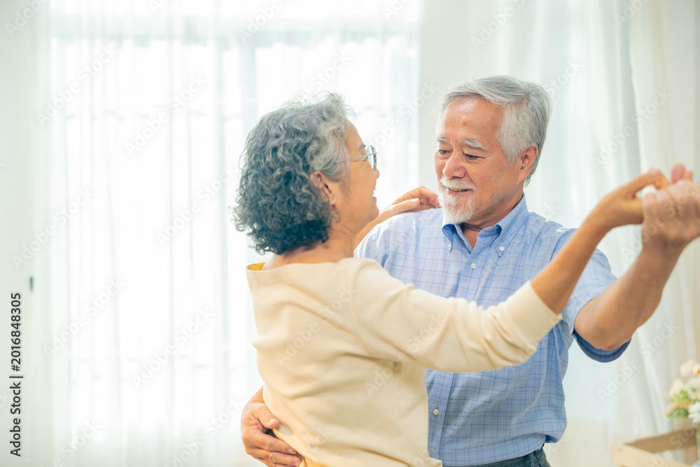 Obraz premium Happy Asian senior couple dancing together at home, elderly husband and wife enjoy active retirement lifestyle indoors, loving older people spending time together, healthy aging joyful relationship.