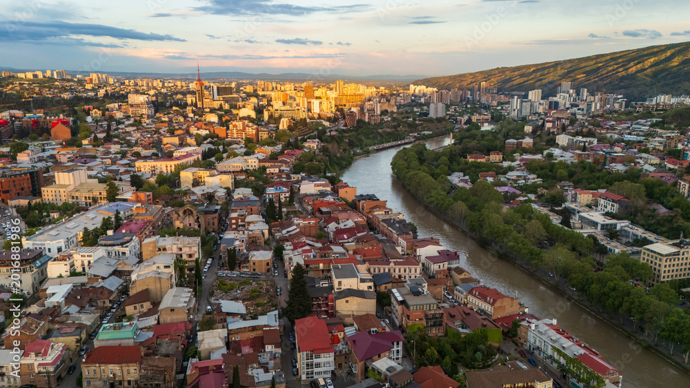 Obraz premium Breathtaking aerial sunset over Tbilisi, Georgia, showcasing the urban sprawl and red rooftops of the old city districts bathed in intense golden hour light with mountain silhouettes
