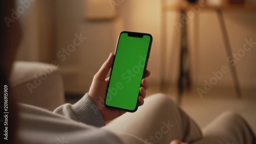 Close-up of a person holding a green screen smartphone while sitting on a couch in a cozy living room with warm lighting.