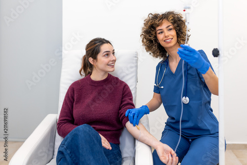 IV drip infusion. Medical treatment, Patient and nurse, Healthcare and Medicine concept. Attractive female nurse adjusting a saline bag for a young patient sitting in a chair at a diagnostic clinic.