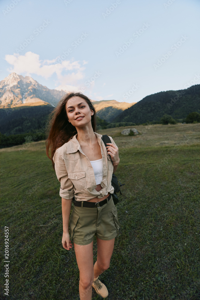 Obraz premium Young woman trekking in an open meadow with distant mountains, wearing a beige jacket and shorts. Natural light highlights a casual lifestyle outdoor moment of exploration.
