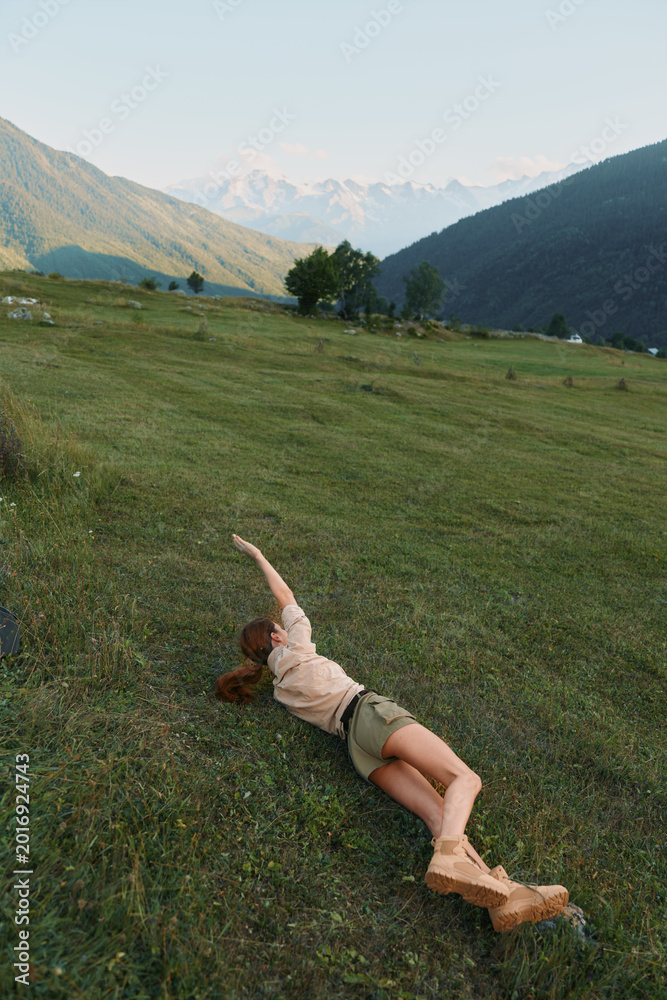 Naklejka premium Young woman relaxing on a hillside meadow with mountains in the background, stretching her arm, casual outdoor lifestyle scene suitable for stock, bright atmosphere, natural light