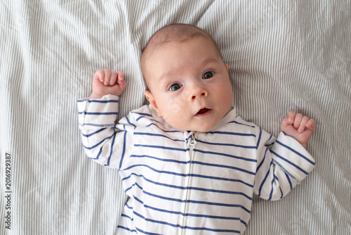 A cute little boy in a striped jumpsuit is lying on a bed with a white striped sheet. Baby, 3 months old. Newborn. Close-up portrait.