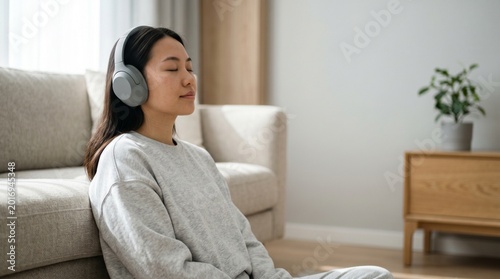 Young Asian woman meditating with headphones while sitting on living room floor.