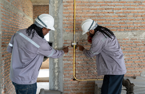 Schilderij op canvas Two Electrician engineer in gray uniform and protective hardhat inspect electrical system in wire PVC pipe using laptop computer and walkie-talkie, especially for security purpose