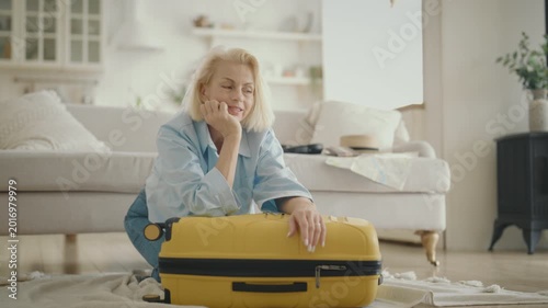 Happy and tired blonde woman closing her big yellow suitcase before traveling. Portrait of single female traveler at home, senior lady dreaming about merry summer vacation on seashore or islands