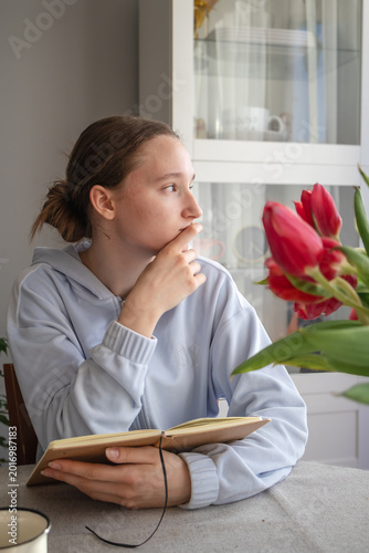 Photos Pensive teenage girl looks away, distracted from reading a book at table near tulips  in warm sunlight
