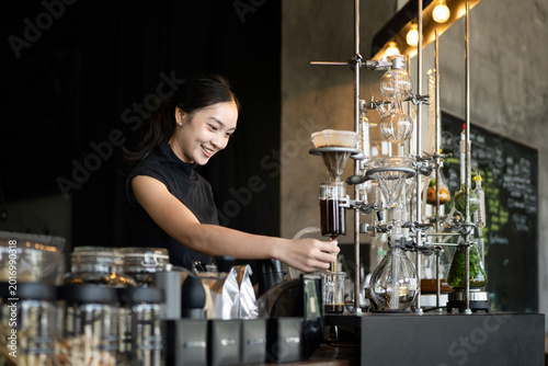 Schilderij op canvas Young Asian woman barista smiling while operating an elegant siphon coffee brewi