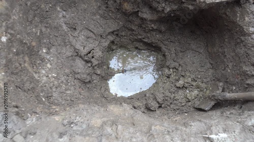 Water main break and repair works in a muddy trench, A deep hole in muddy soil during emergency water pipe repairs. Water sprays under pressure from a ruptured underground pipe, forming a puddle.