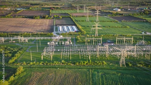 Aerial tracking shot from left to right of electrical substation with a large-scale battery energy storage system (BESS), high-voltage pylons and power lines in the background. 
