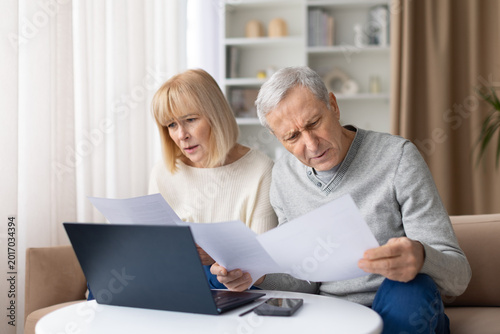Fotografie Concerned senior couple reading financial papers at home, looking worried about